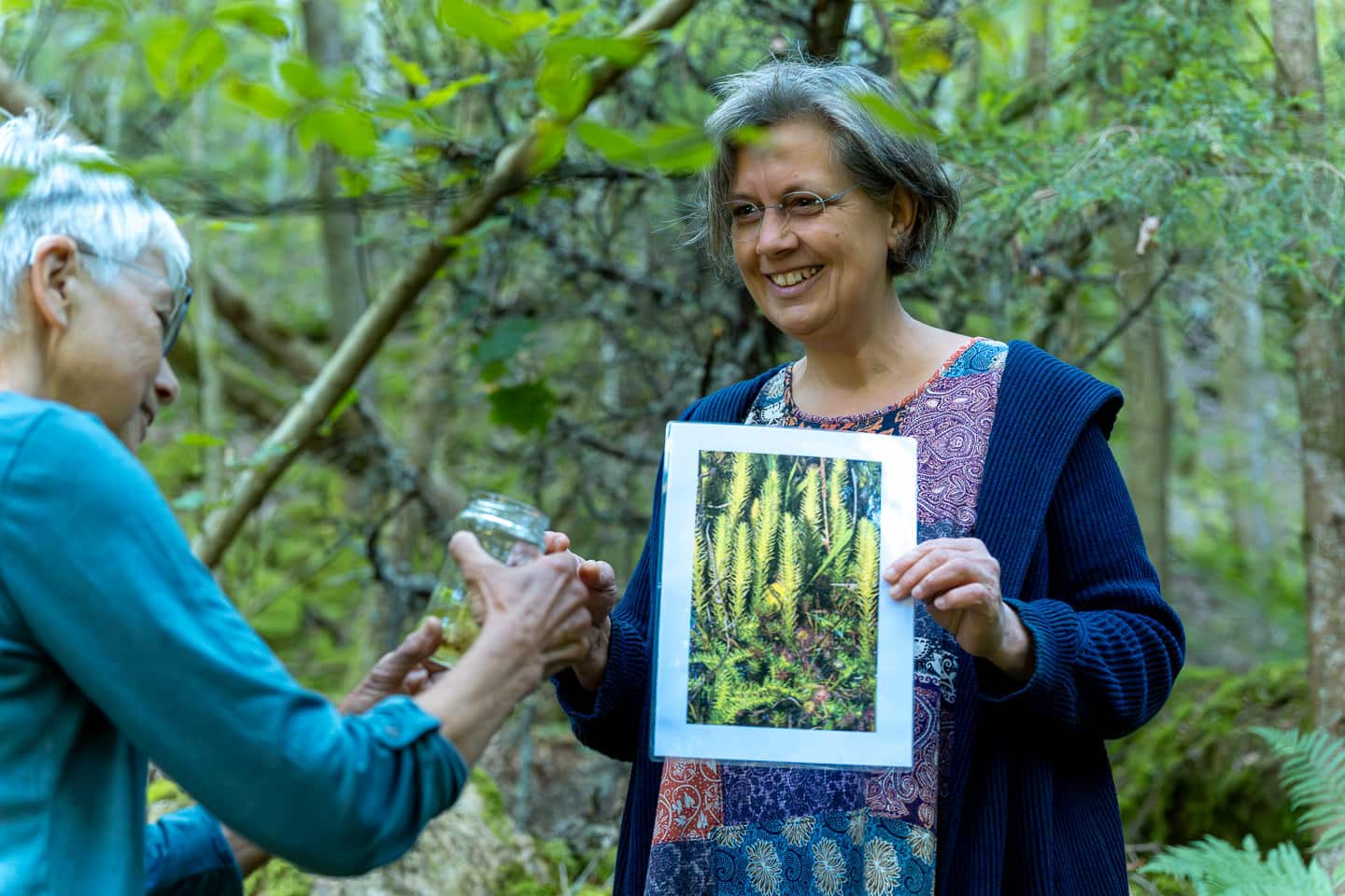 Eine Frau hält ein Bild mit einer Pflanze und zeigt einer anderen Frau etwas in einem Schraubglas