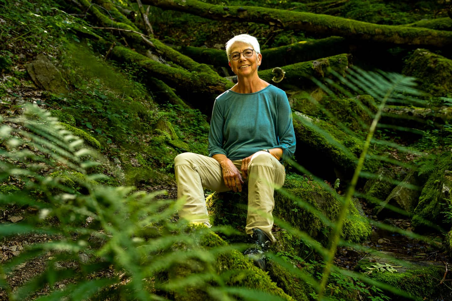 Eine Frau mit kurzen weißen Haaren sitzt zwischen Farn und bemoosten Baumstämmen