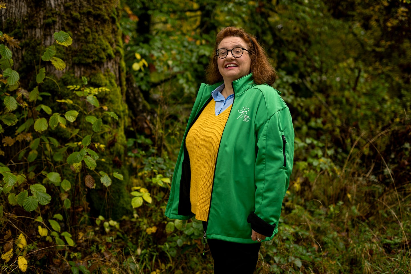 Eine Frau mit roten Haaren und grüner Regenjacke und Brille im Wald