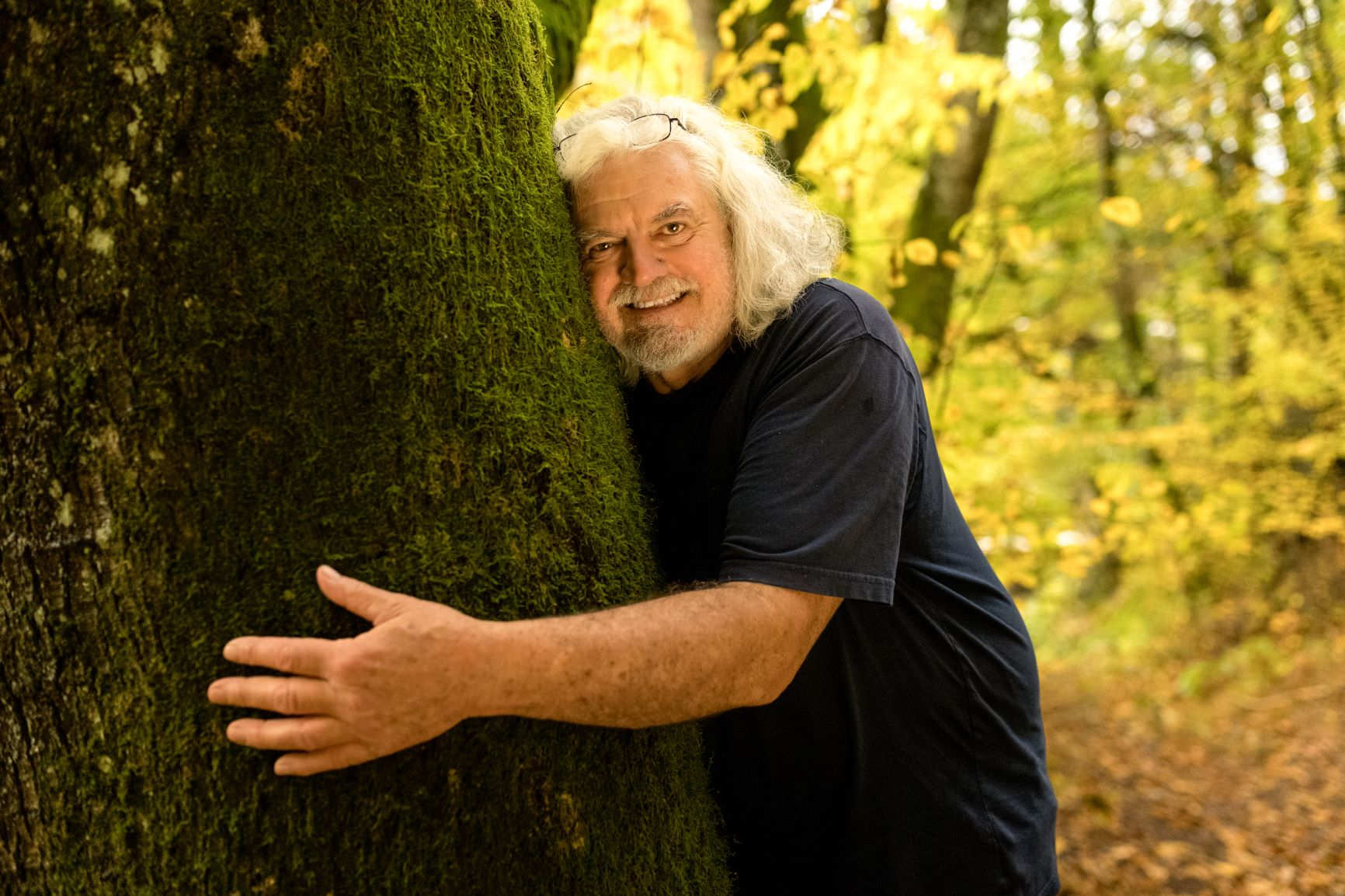 Ein Mann mit langen weißen Haaren umarmt einen Baum